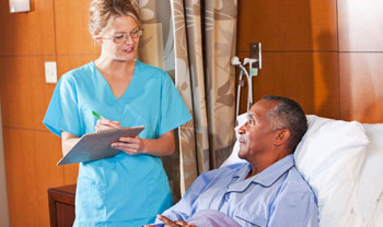 Female nurse holding a clip board and writing, looking at patient who is in bed standing 